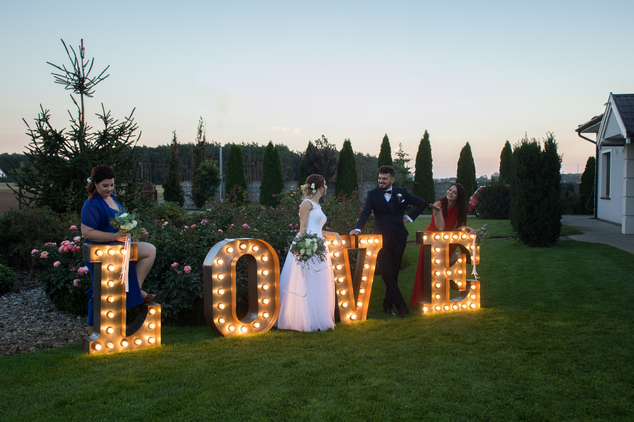 Para młoda pozuje przy podświetlanym napisie LOVE, otoczona druhnami w ogrodzie z różami, podczas zachodu słońca.