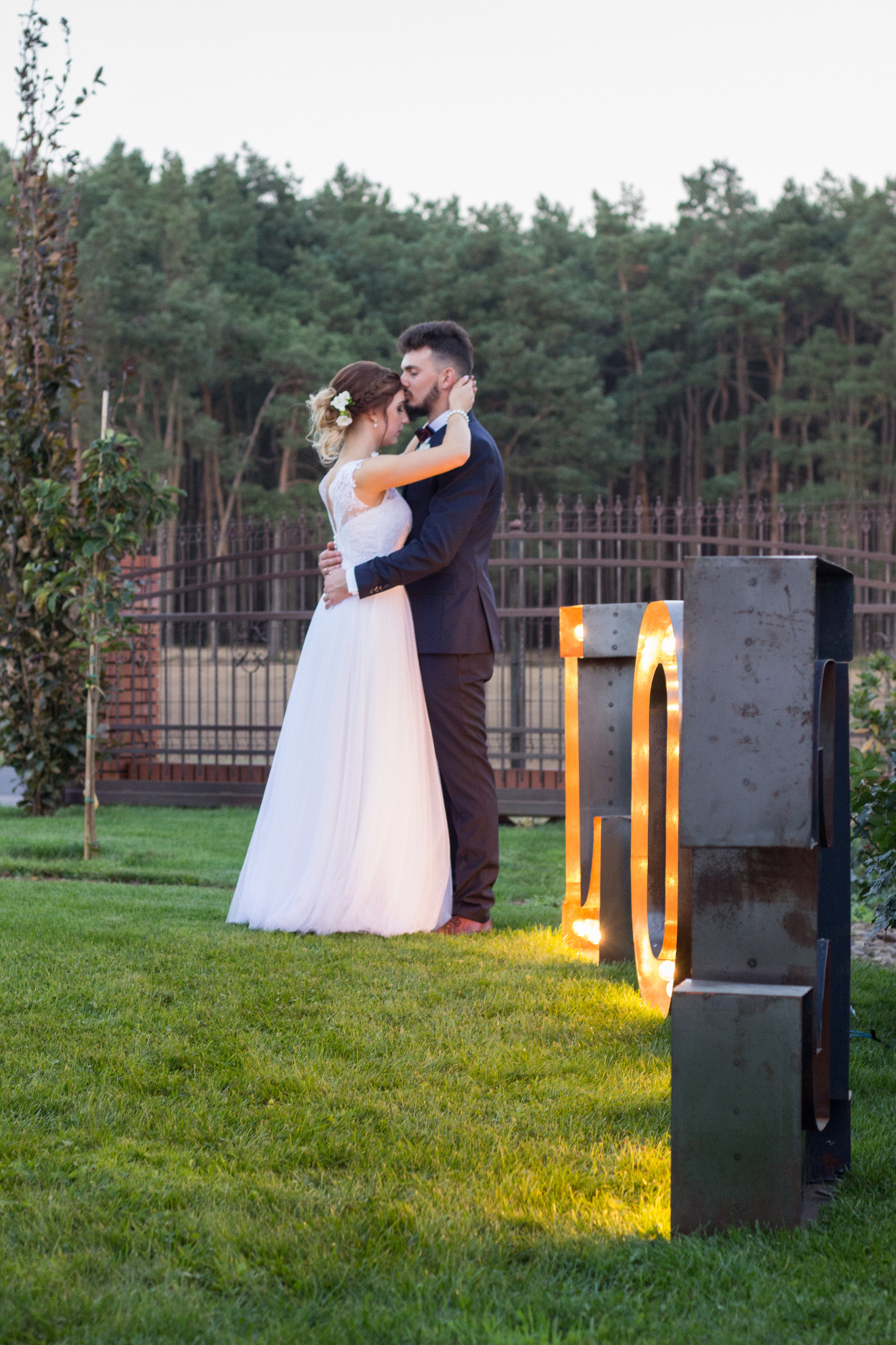 Para młoda w objęciach na trawniku przed podświetlanymi literami, panna młoda w białej sukni z koronkową górą, pan młody w granatowym garniturze, na tle zieleni i ogrodzenia.