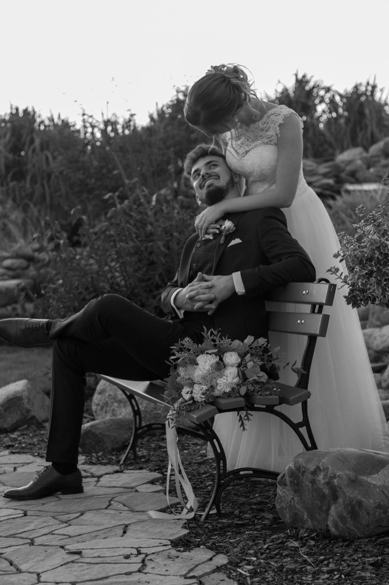 Czarno-białe zdjęcie pary młodej: panna młoda obejmuje pana młodego siedzącego na ławce w ogrodzie, bukiet ślubny leży obok.