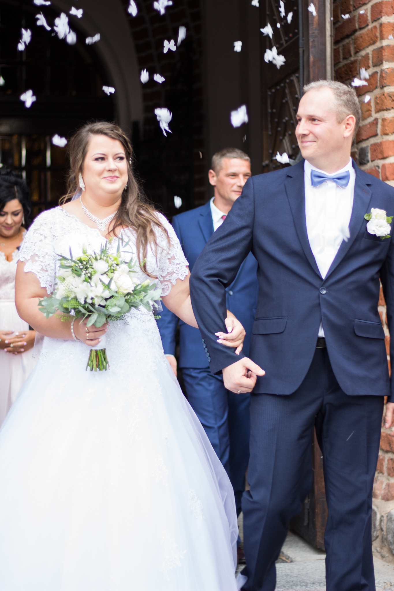 Para młoda wychodzi z kościoła, obsypywana białymi płatkami konfetti, panna młoda w białej sukni z koronkową górą i bukietem, pan młody w granatowym garniturze z muchą, uśmiechnięci.