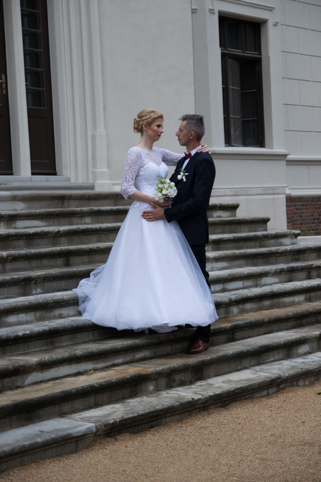 Para młoda na kamiennych schodach przed budynkiem z kolumnami, panna młoda w sukni z koronkowymi rękawami trzyma bukiet białych kwiatów, pan młody w czarnym garniturze z bordową muchą.