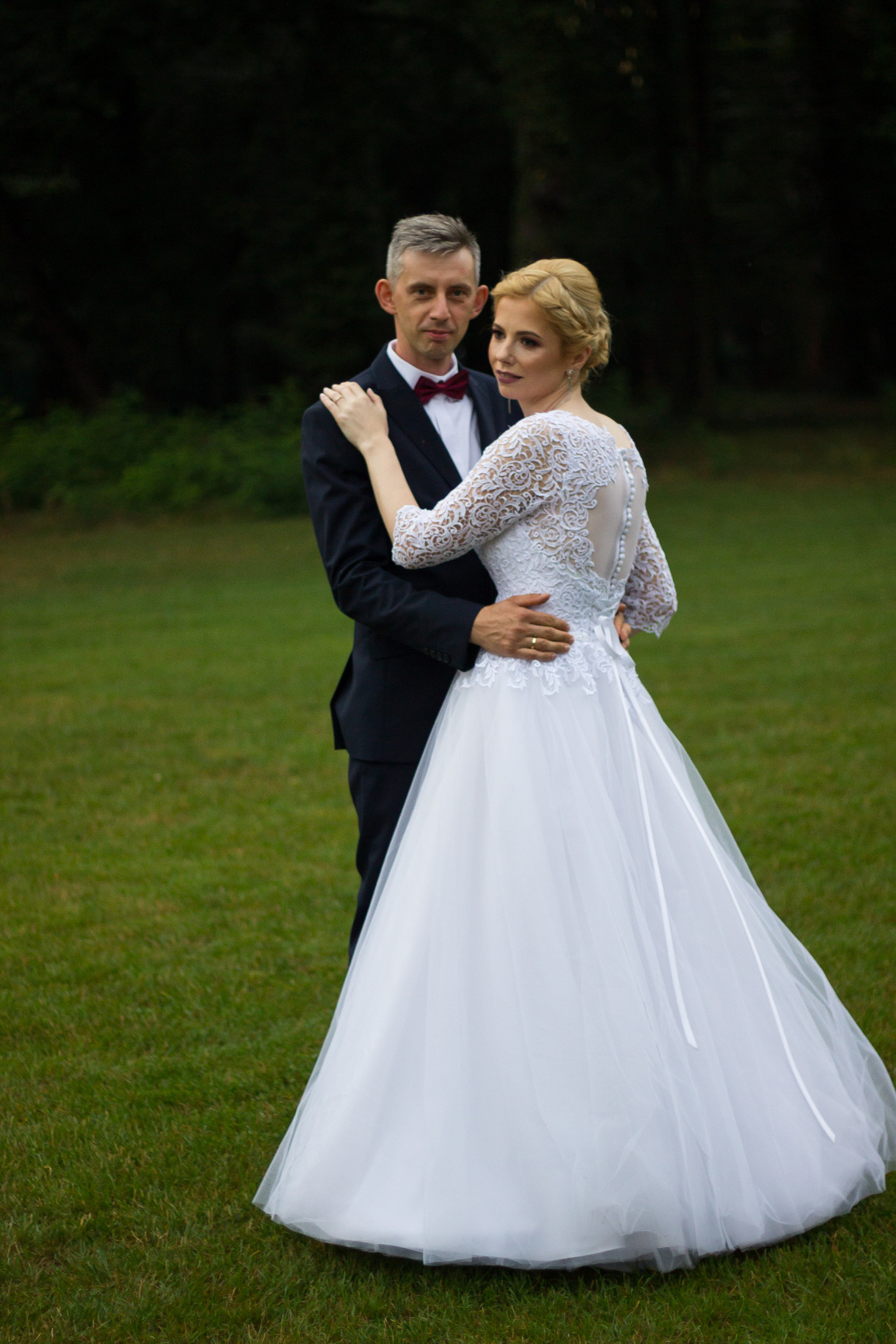 Para młoda pozuje na zielonej łące: panna młoda w koronkowej sukni z odkrytymi plecami, pan młody w granatowym garniturze z bordową muchą.