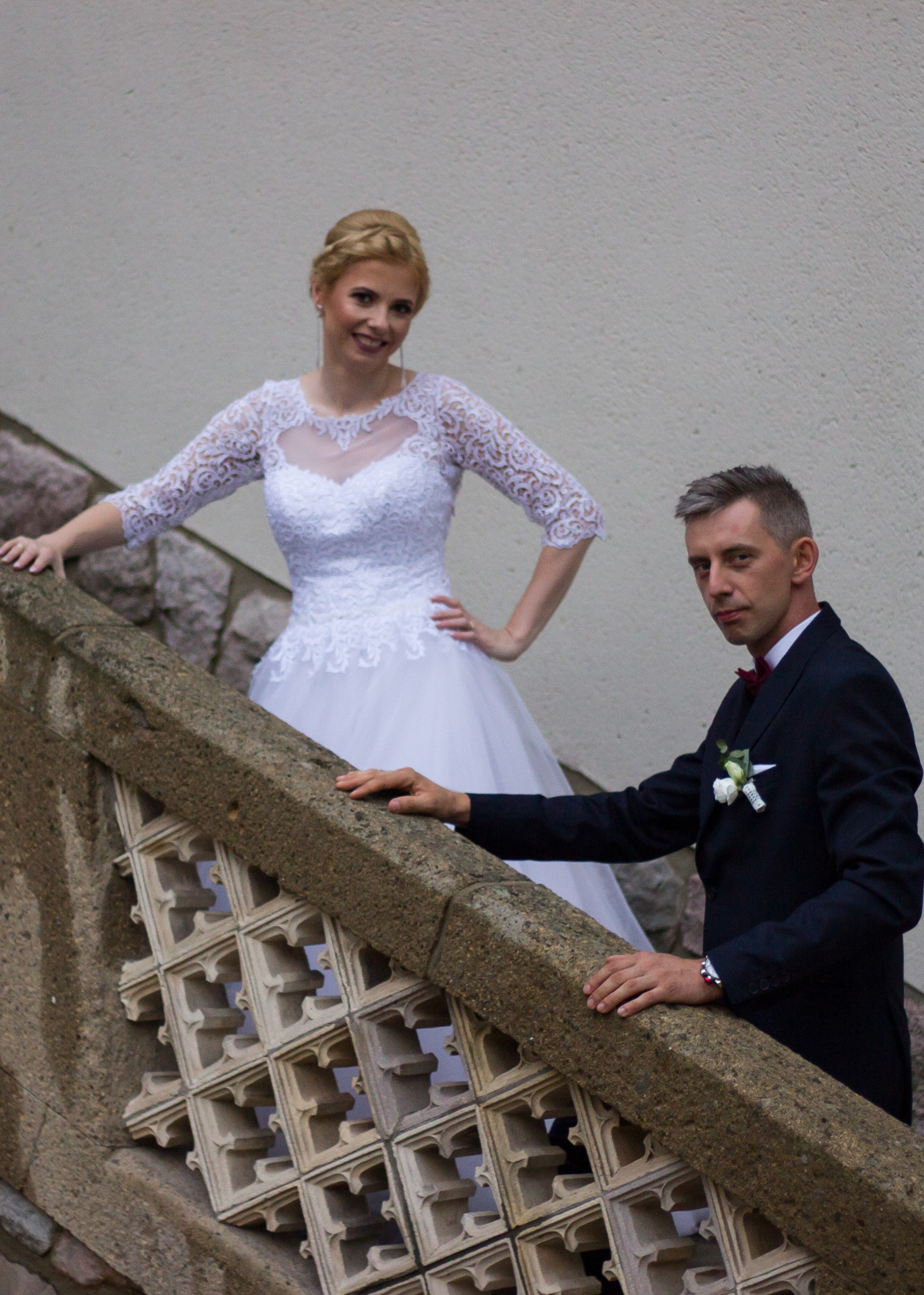 Para młoda pozuje na schodach z ozdobną kamienną balustradą; panna młoda w koronkowej sukni z długim rękawem, pan młody w granatowym garniturze z bordową muchą.