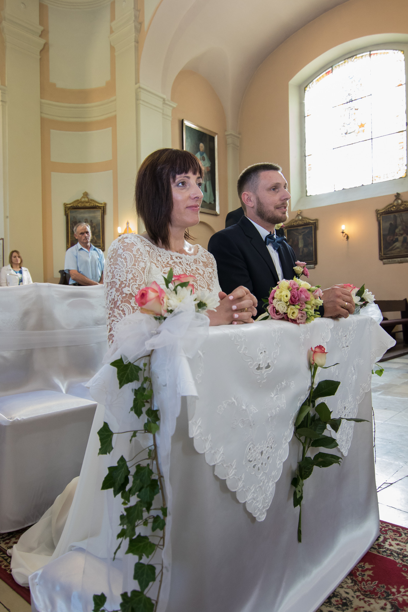 Para młoda siedzi w kościele podczas ceremonii ślubnej, z bukietami kwiatów i dekoracjami z bluszczu na klęczniku, w tle widoczni goście i obrazy.