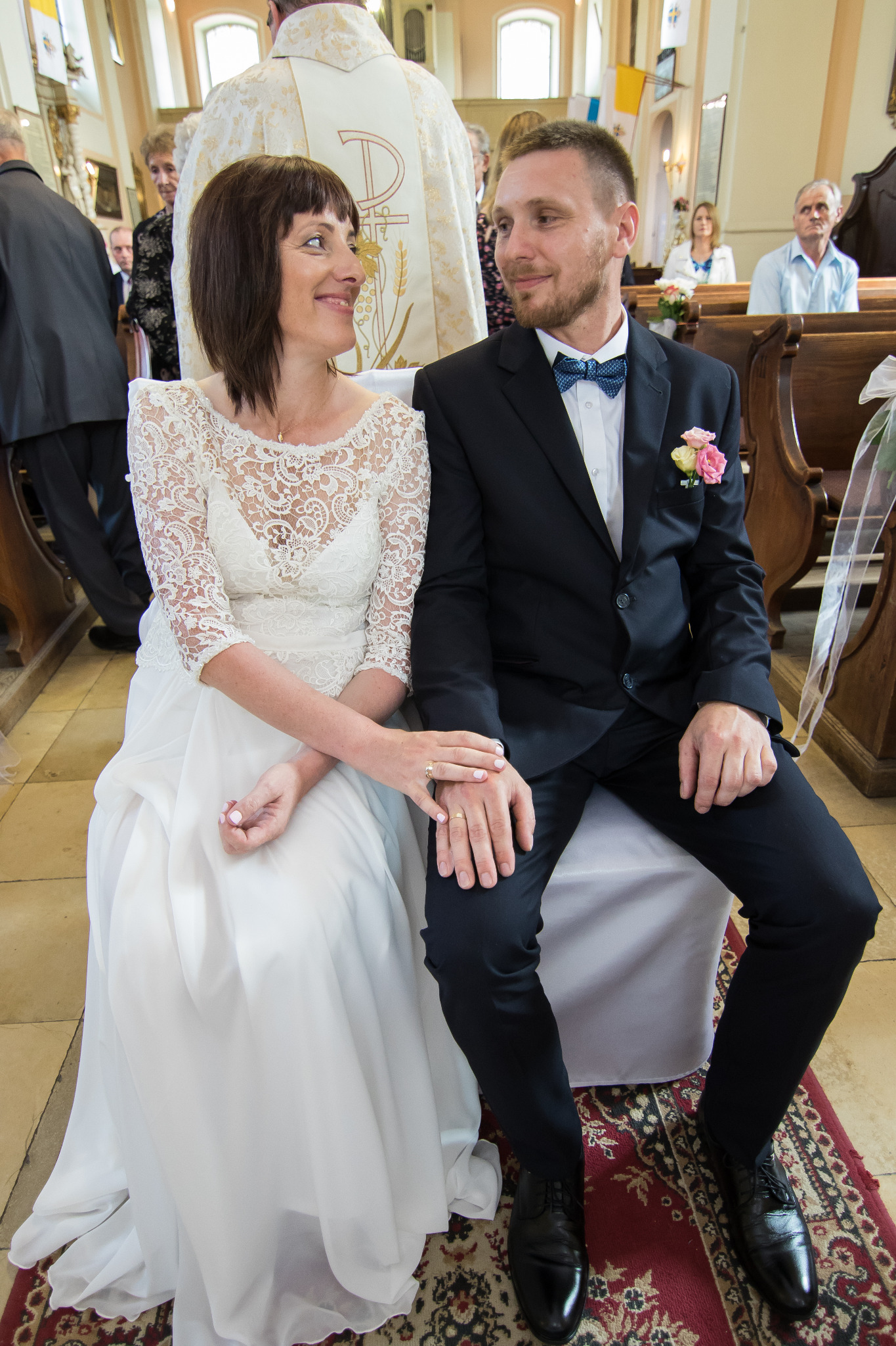 Para młoda siedzi obok siebie w kościele, trzymając się za ręce, z uśmiechem patrząc na siebie podczas ceremonii ślubnej.