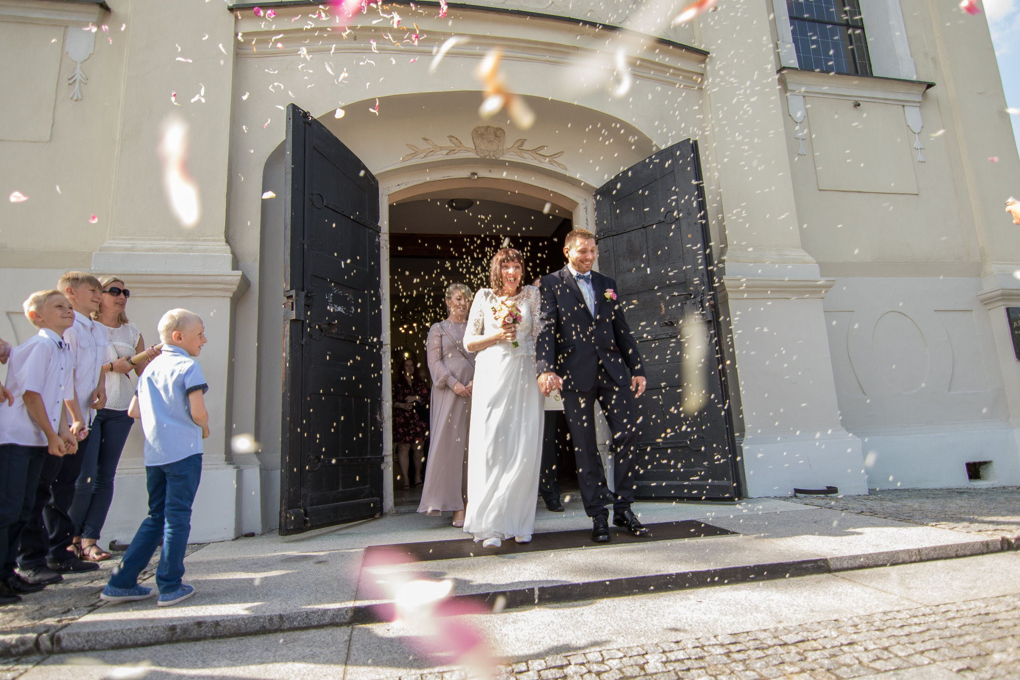 Para młoda wychodzi z kościoła, obsypywana konfetti, w tle goście weselni i elewacja budynku.