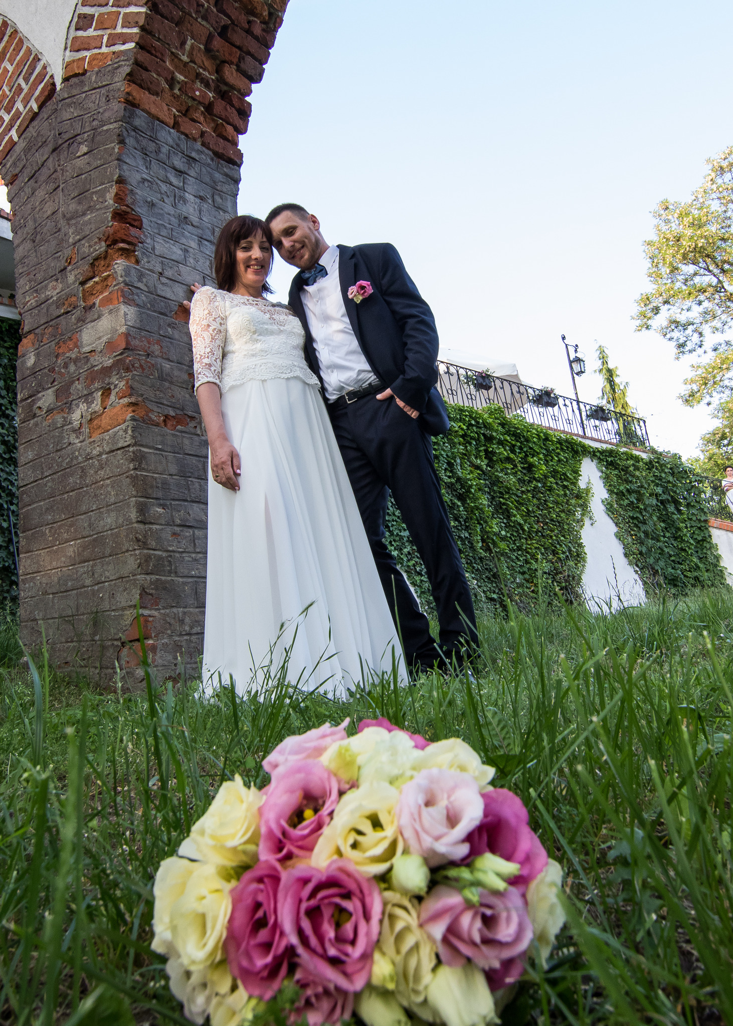 Para młoda pozuje w plenerze przy ceglanym łuku, bukiet kwiatów w pierwszym planie na trawie.