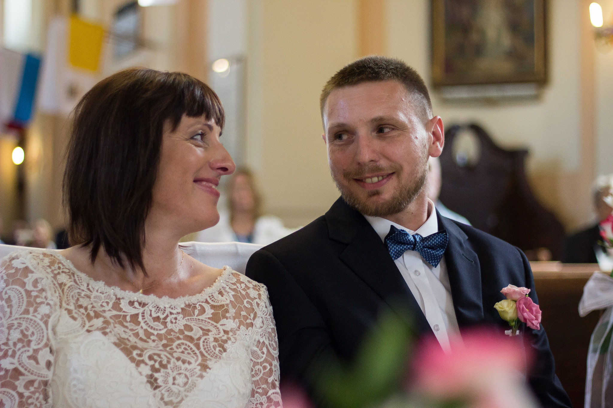 Para młoda siedzi obok siebie, spoglądając na siebie z uśmiechem podczas ceremonii ślubnej w kościele, widoczny bukiecik w butonierce pana młodego.