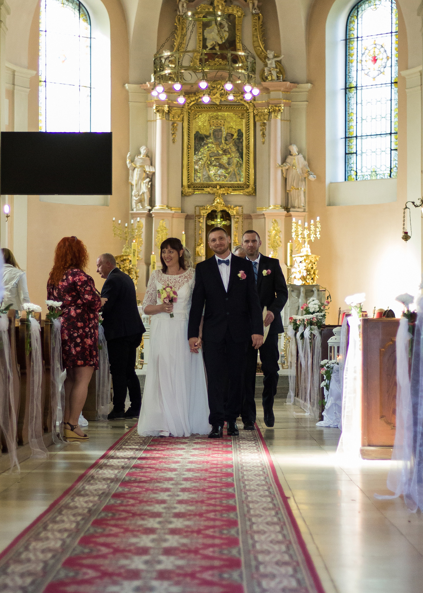 Para młoda w kościele idzie wzdłuż czerwonego dywanu, za nimi drużba, w tle ołtarz i goście.