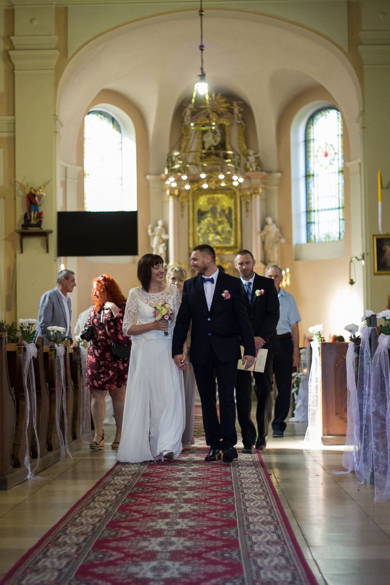 Para młoda, uśmiechnięta, wychodzi z kościoła po ceremonii ślubnej, idąc po czerwonym dywanie, za nimi goście.