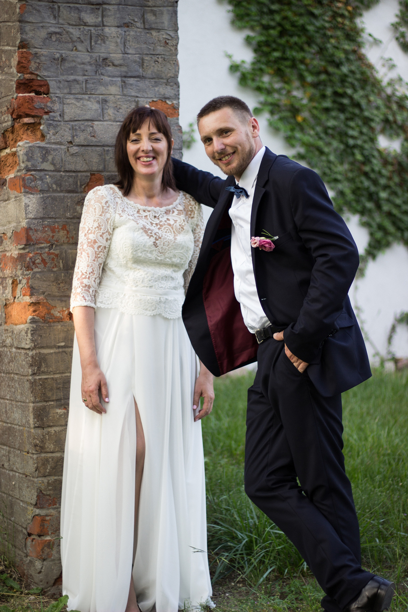 Uśmiechnięta para młoda pozuje przy ceglanym filarze porośniętym bluszczem: Panna młoda w koronkowej sukni z rozcięciem, pan młody w granatowym garniturze z muszką i butonierką.