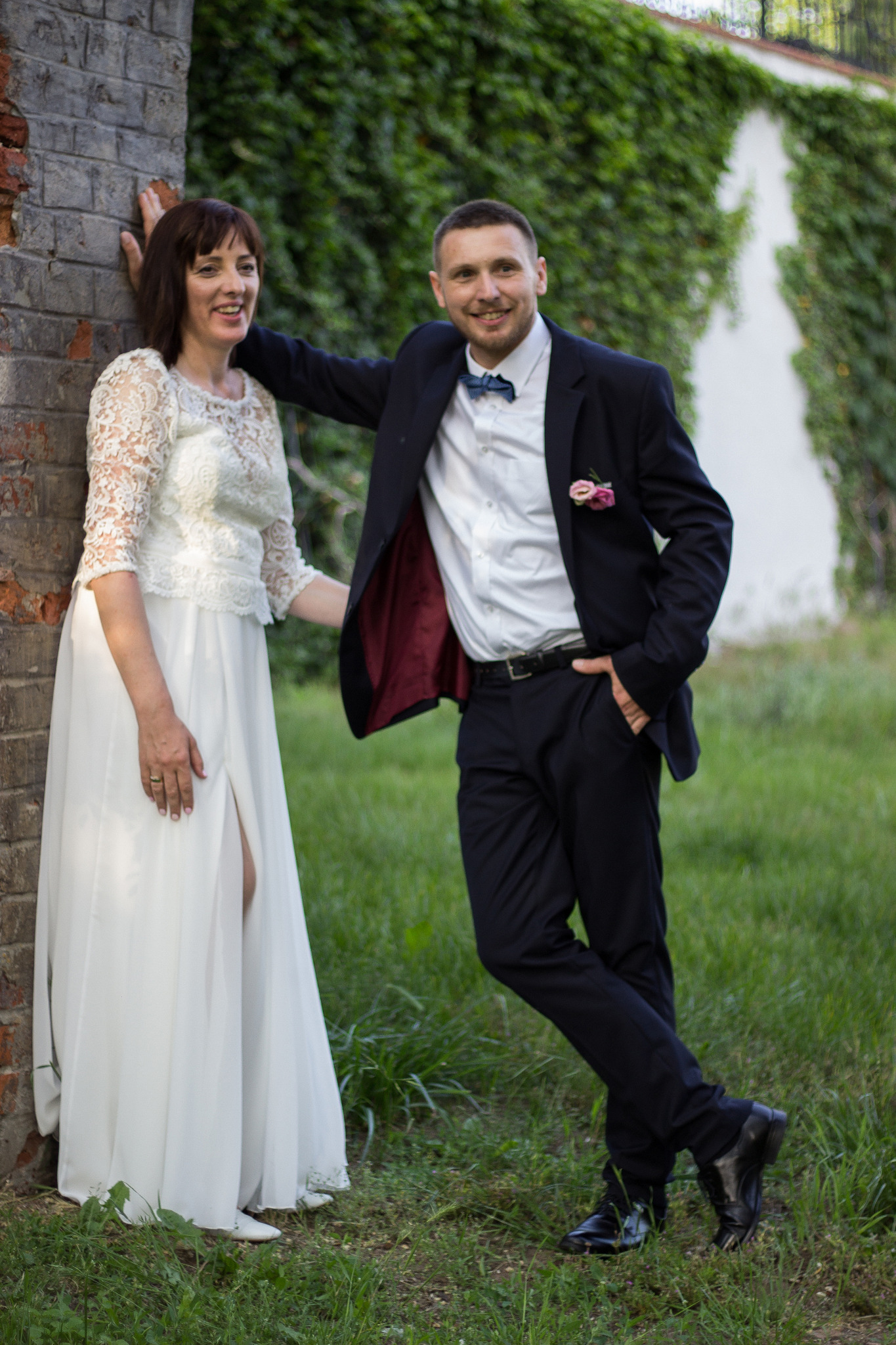 Para młoda: kobieta w białej sukni z koronkową górą i rozcięciem, mężczyzna w granatowym garniturze z muchą, opierają się o ceglany filar na tle zielonej ściany z bluszczu.