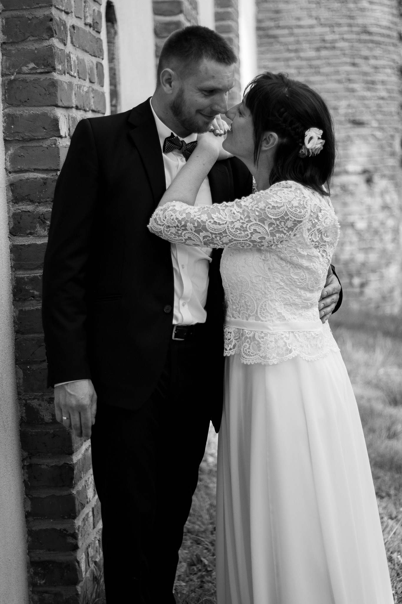 Czarno-białe zdjęcie pary młodej: panna młoda w koronkowej sukni dotyka twarzy pana młodego w muszce, stoją przy ceglanej ścianie.