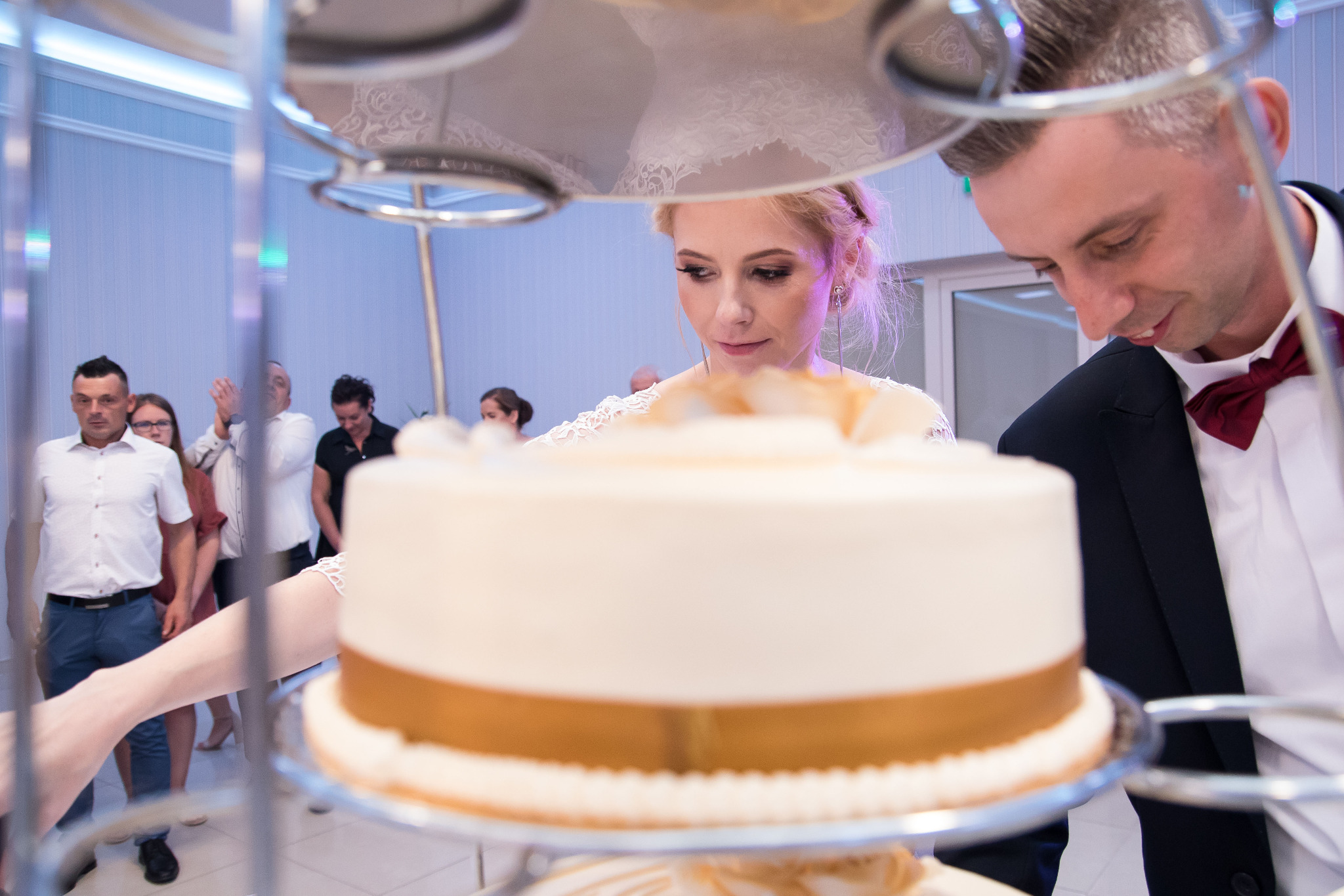 Panna młoda i pan młody kroją tort weselny z kremowym lukrem i złotym paskiem, goście w tle.