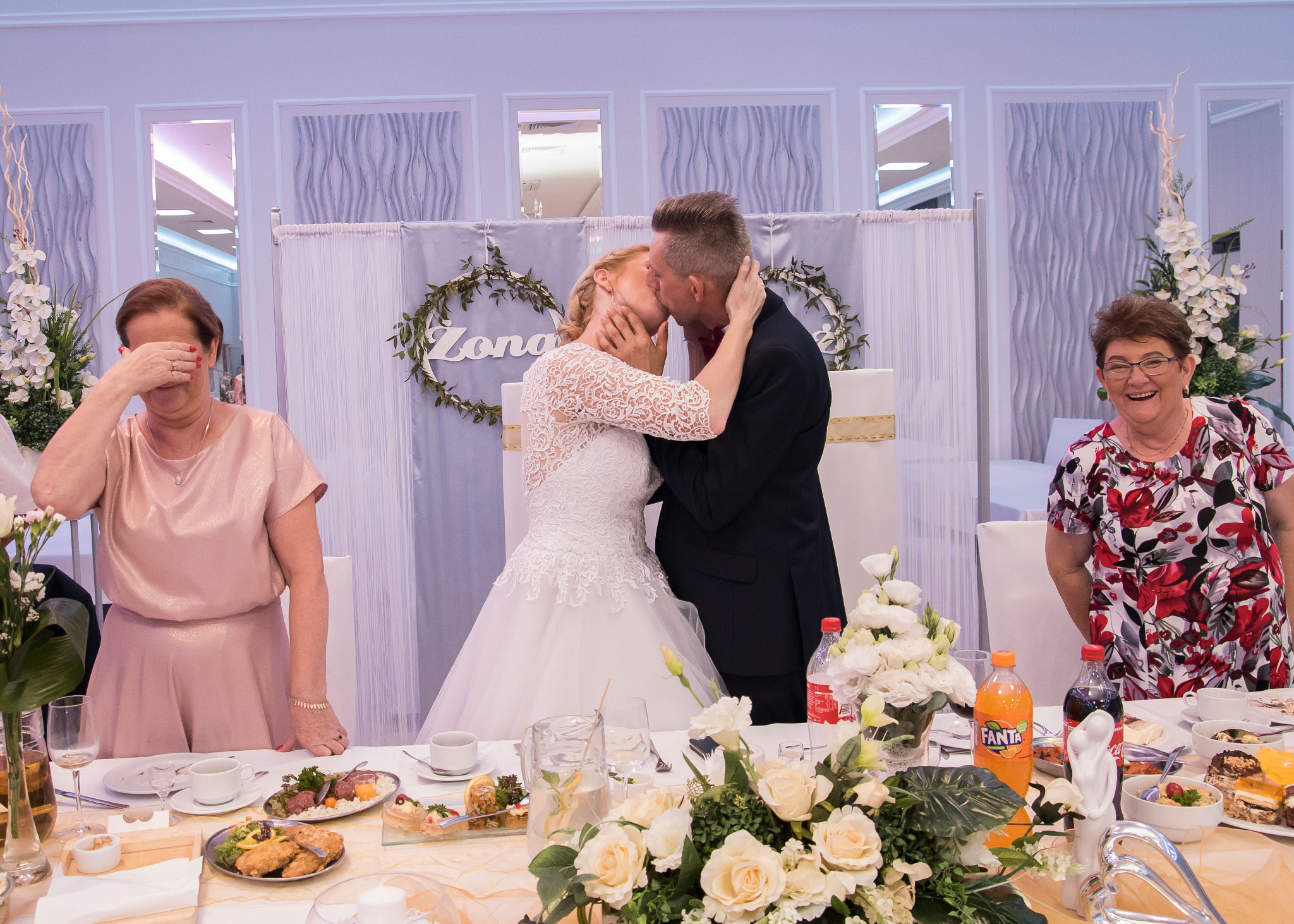 Pocałunek pary młodej na tle dekoracji z napisem Żona, wzruszona kobieta z zakrytymi oczami i uśmiechnięta kobieta w kwiecistej bluzce przy stole z jedzeniem i napojami.