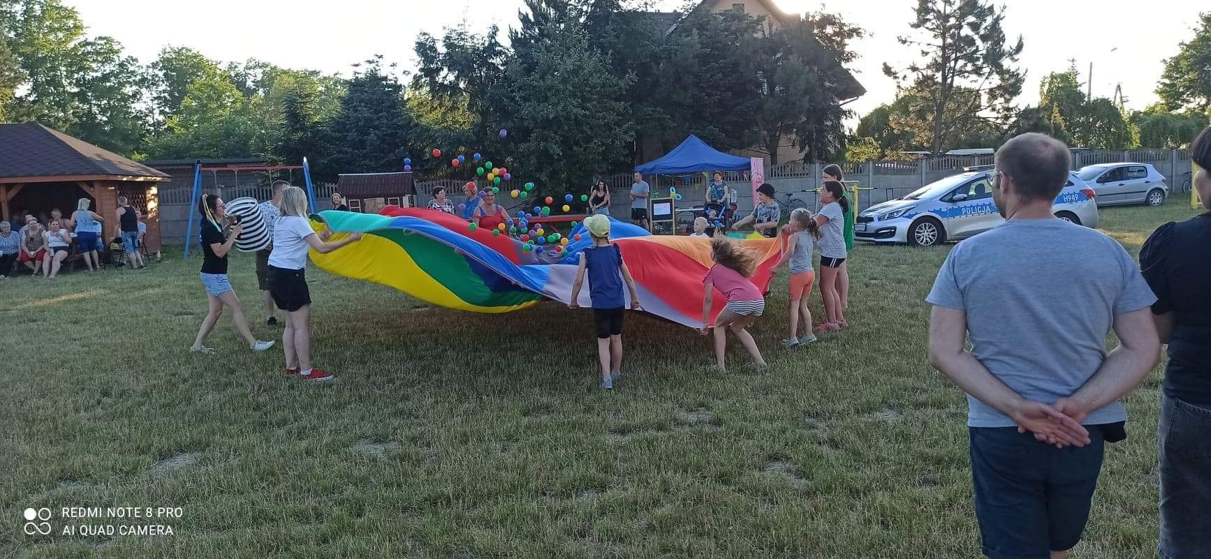 Grupa dzieci i dorosłych trzymających kolorową chustę animacyjną wypełnioną piłeczkami na trawniku podczas imprezy plenerowej, w tle widoczna altana, namiot i samochody.