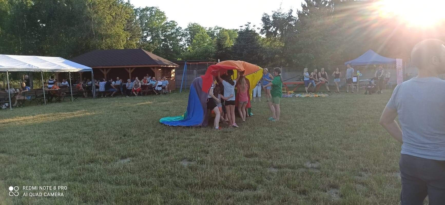 Grupa dzieci bawi się pod wielokolorową chustą animacyjną na trawniku podczas imprezy plenerowej, w tle widoczne namioty i siedzący ludzie.
