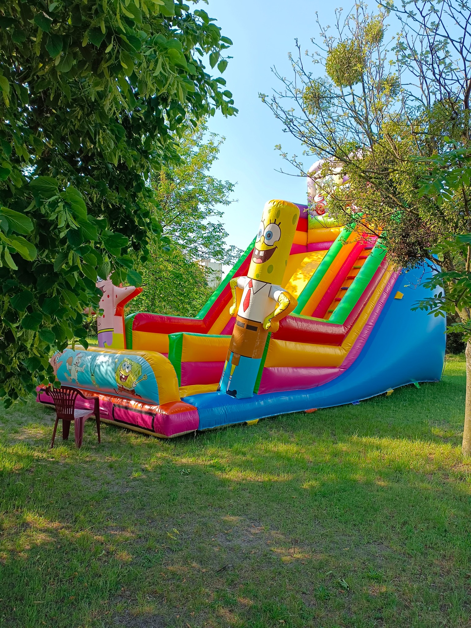 Kolorowy dmuchany zamek ze zjeżdżalnią w kształcie SpongeBoba na trawniku, widoczne drzewa i krzesło obok.