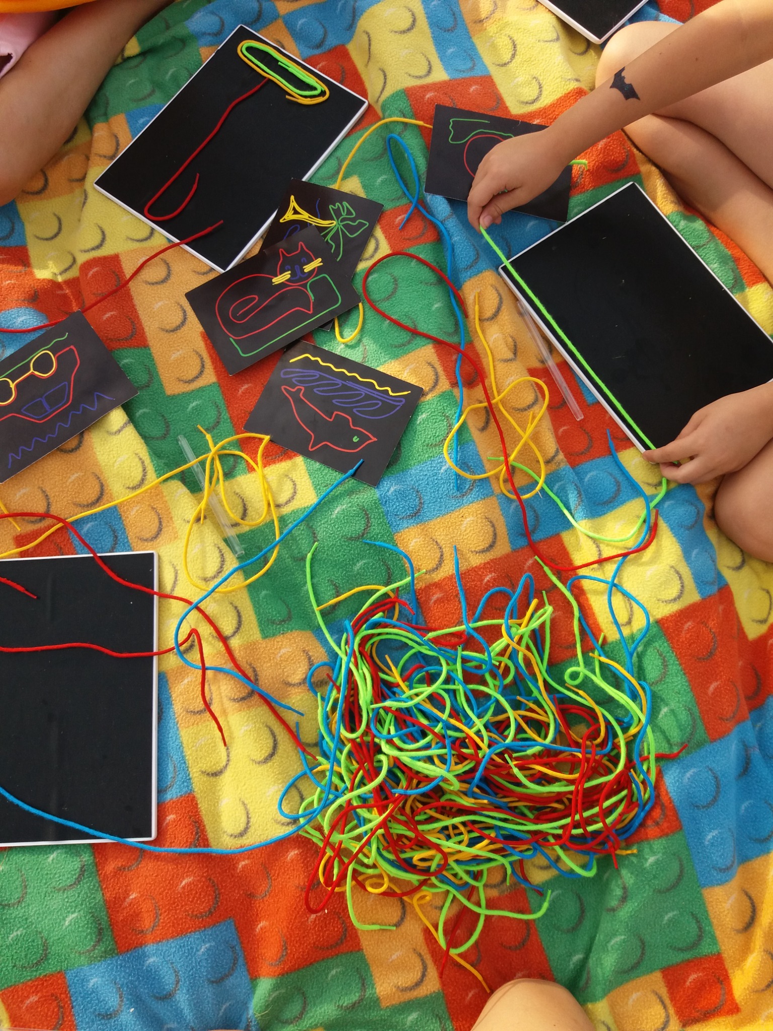 Dzieci bawią się kolorowymi sznurkami i czarnymi tablicami z narysowanymi wzorami na kocu w kształcie klocków Lego.