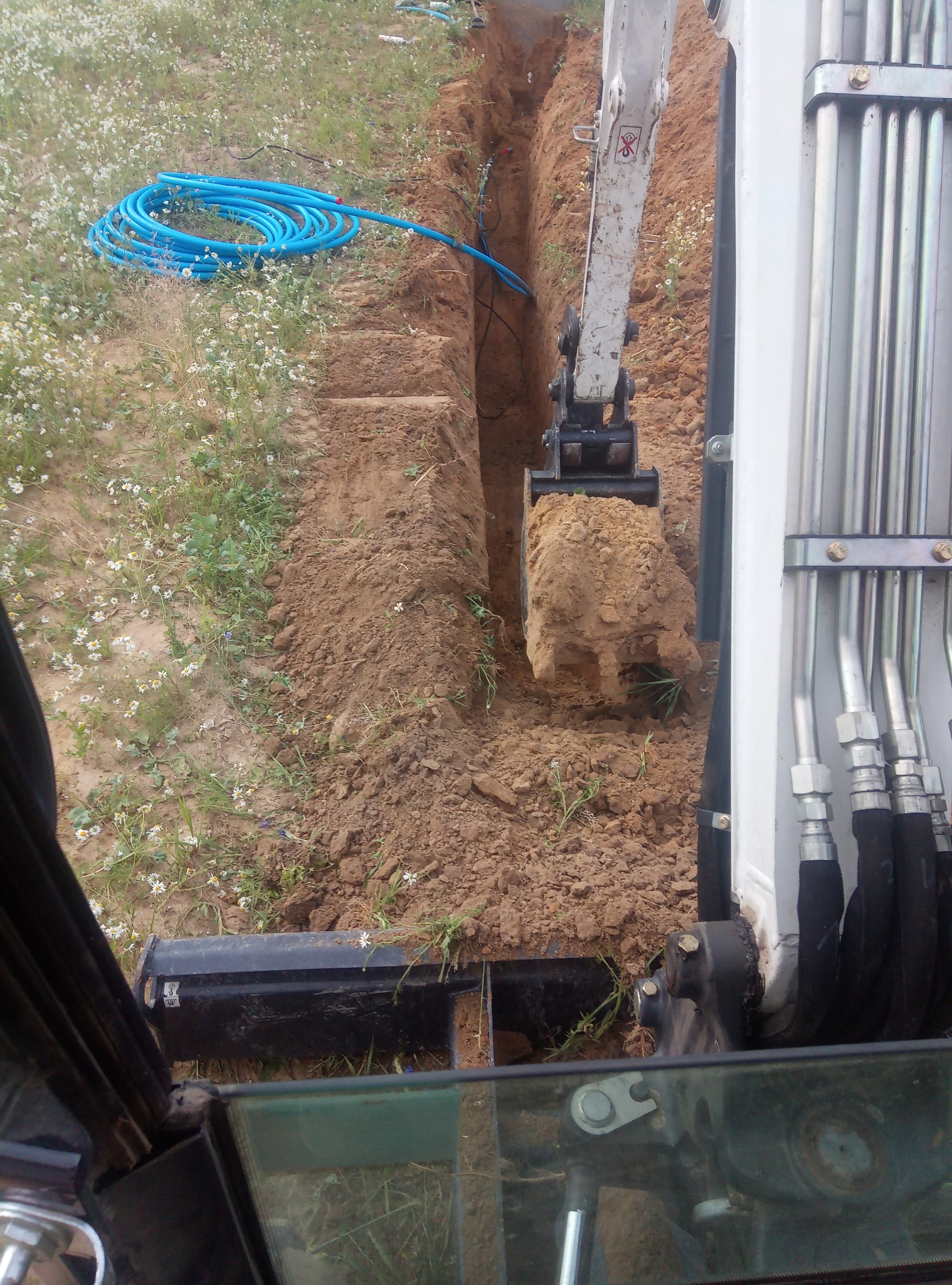 Mini koparka w trakcie wykonywania wąskiego wykopu pod instalacje w Krasnem, obok leży zwój niebieskich rur.