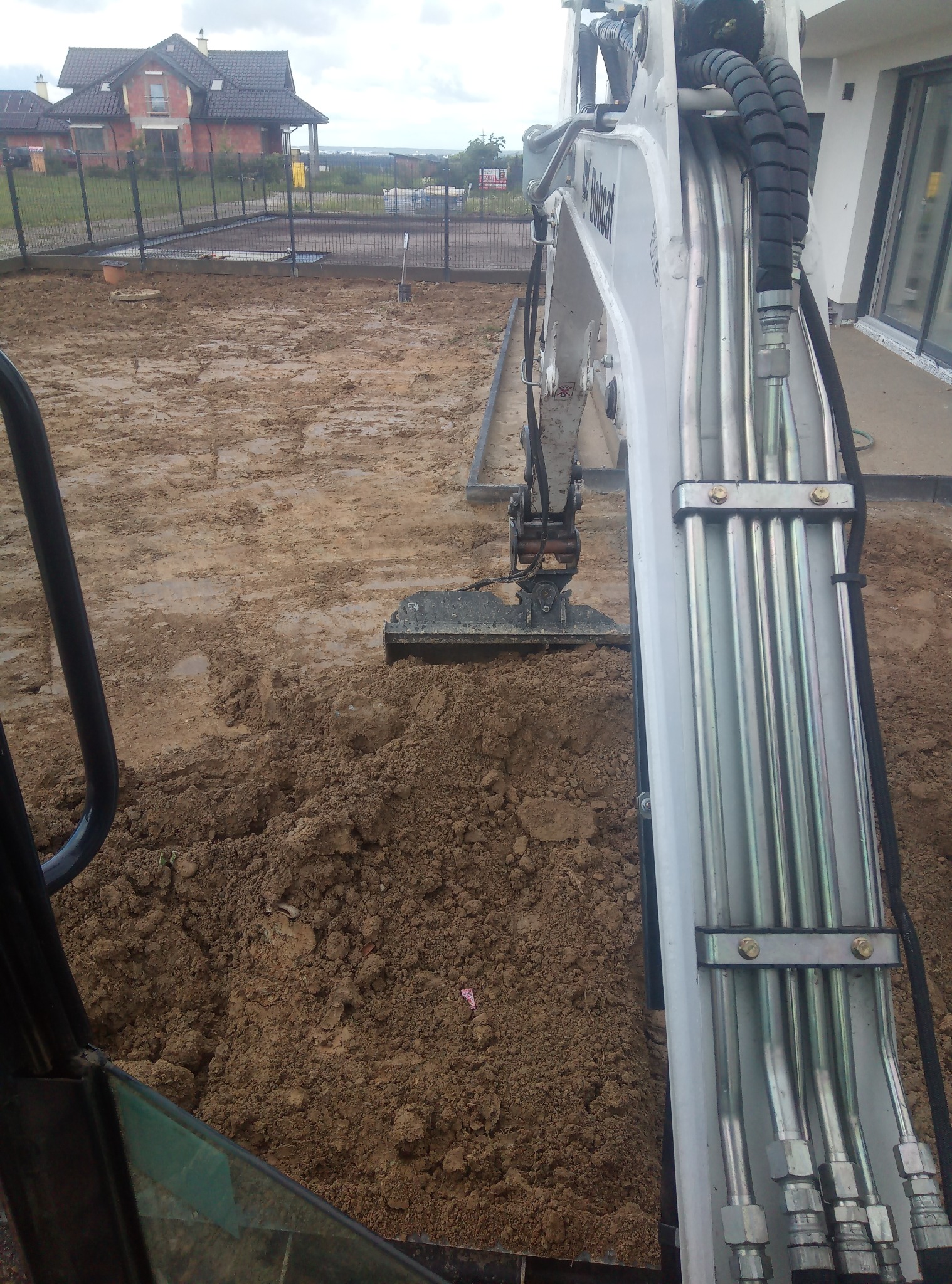 Widok z kabiny małej koparki Bobcat na wykopaną ziemię, w tle ogrodzony teren budowy z domem jednorodzinnym.