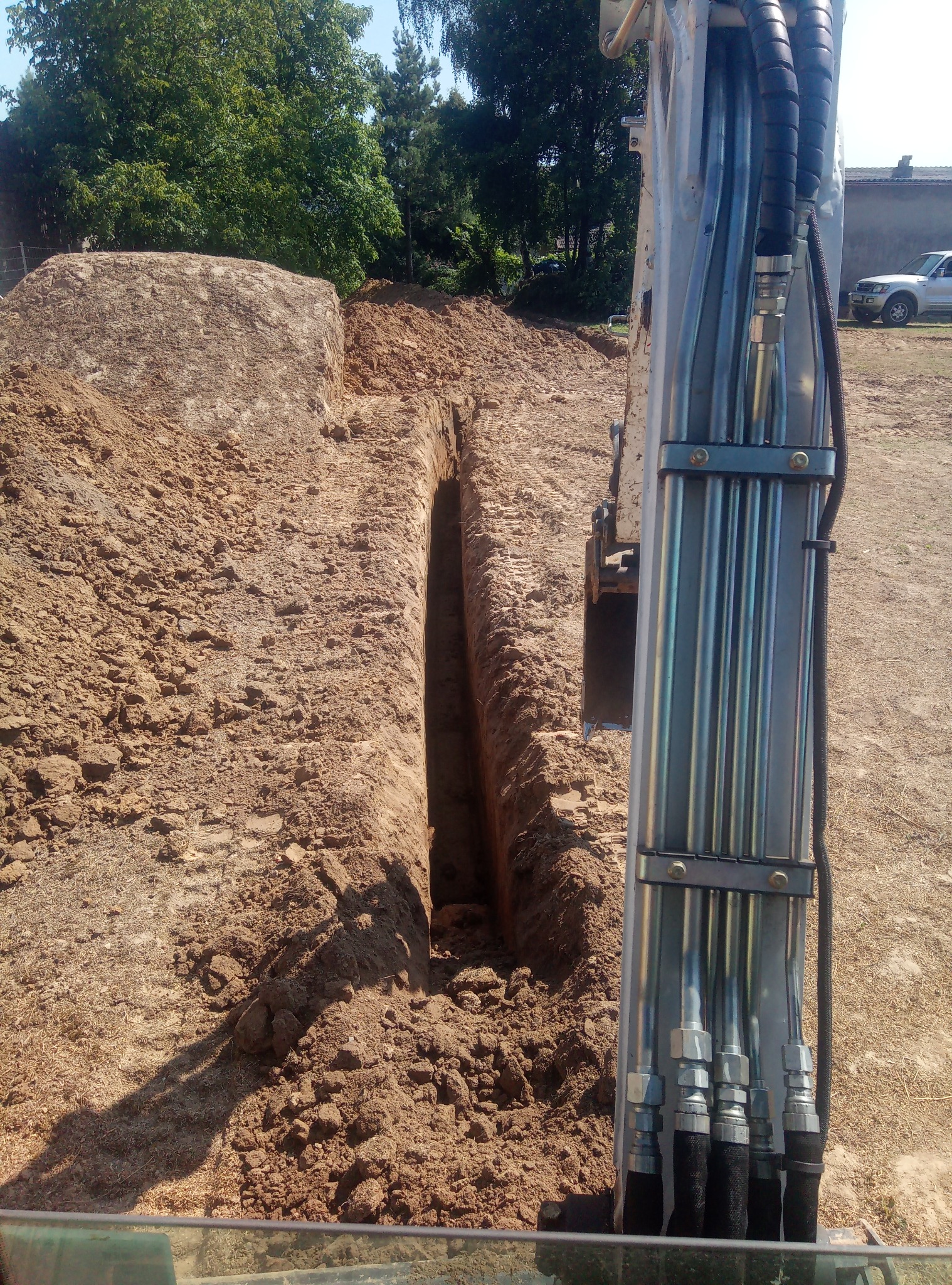 Wąski wykop w piaszczystej glebie, widok z kabiny koparki na ramię robocze z przewodami hydraulicznymi, usypana pryzma ziemi w tle.