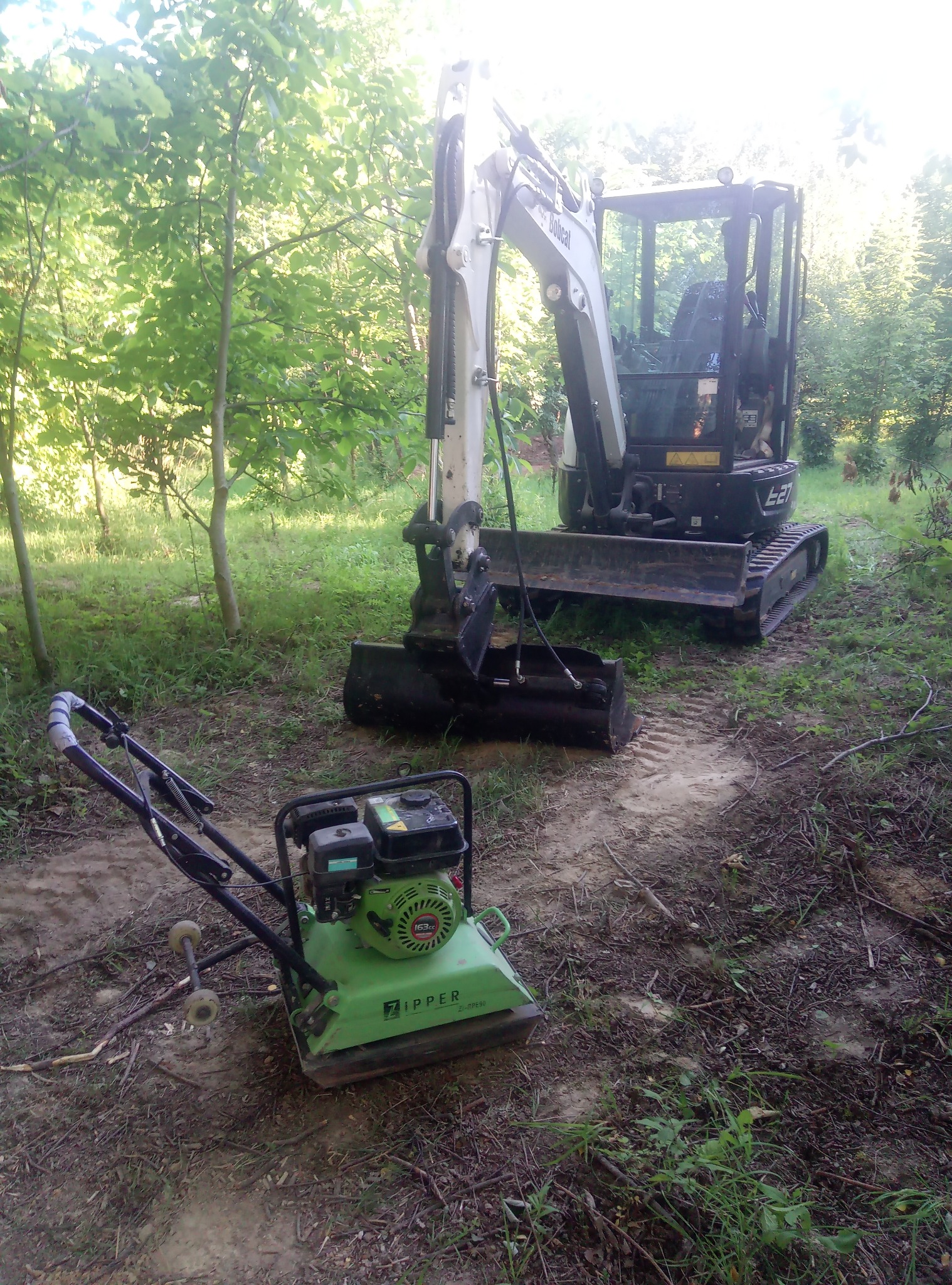 Mini koparka Bobcat E27 i zagęszczarka gruntu Zipper na leśnej działce przed rozpoczęciem prac, widoczne drzewa i trawa.