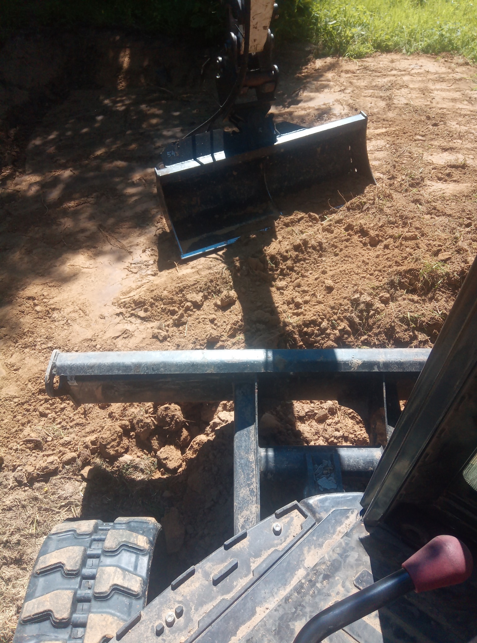 Koparka z łyżką zawieszoną nad nierównym, piaszczystym terenem w słoneczny dzień; widok z perspektywy operatora.