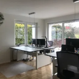 Jasne biuro z biurkami, komputerami i widokiem na zieleń za oknem, oświetlone nowoczesnymi lampami sufitowymi.