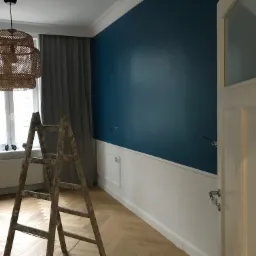 Pomieszczenie w trakcie prac: ściana pomalowana na granatowo od g&oacute;ry, biała boazeria na dole, drewniana drabina malarska na parkiecie w jodełkę, widoczne gniazdka elektryczne bez osłon.