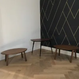 Trzy drewniane stoliki o różnych kształtach na parkiecie ułożonym w jodełkę, na tle białej ściany i ściany z czarną tapetą w złote geometryczne wzory.