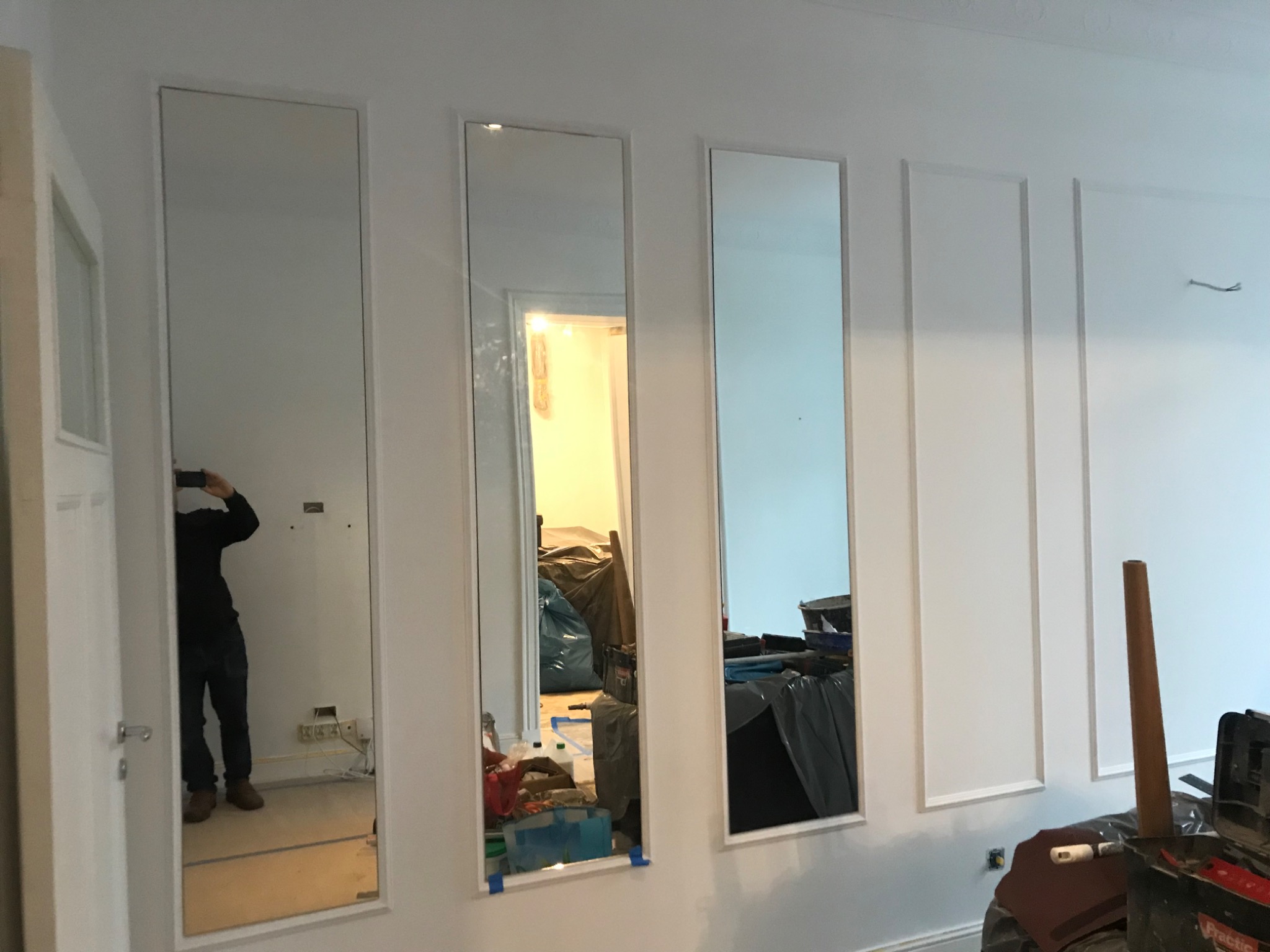 Montaż trzech wysokich luster w wąskich, białych ramach na białej ścianie z dekoracyjnymi listwami; widoczne narzędzia i bałagan w tle.