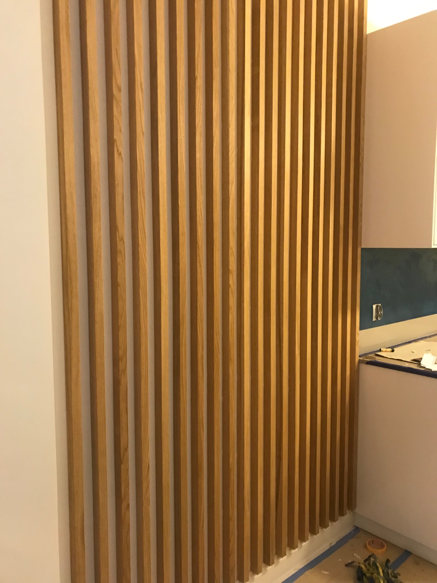 Drewniane lamele pionowe tworzące dekoracyjną ścianę działową, widoczne zabezpieczenia podłogi taśmą malarską, fragment blatu z narzędziami w tle.