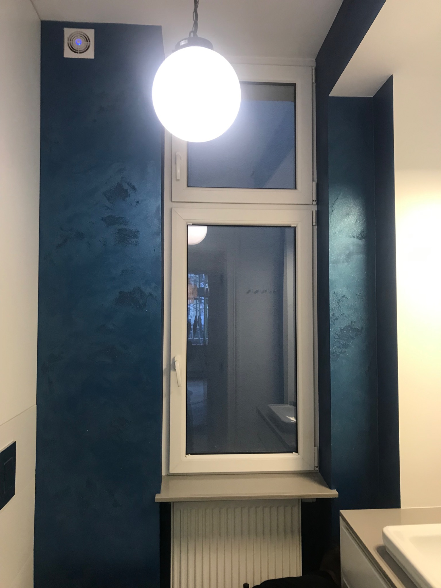 Ściana w odcieniach granatu z dekoracyjną fakturą obok białego okna PCV, oświetlona wiszącą lampą kulą.