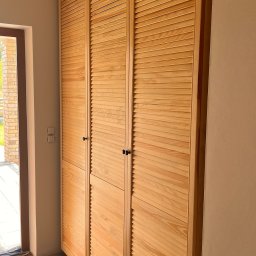 Cool-Ka MEBLE - Trzydrzwiowa szafa wnękowa z jasnego drewna, z lamelowymi frontami i czarnymi gałkami, widziana z boku, z widoczną częścią podłogi zabezpieczonej folią malarską i wejście do budynku w tle.