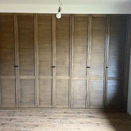 Cool-Ka MEBLE - Duża, drewniana szafa z lamelowymi drzwiami, zajmująca całą ścianę, z widoczną podłogą z paneli imitujących drewno i fragmentem drzwi po lewej stronie.