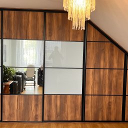 Cool-Ka MEBLE - Szafa wnękowa z przesuwnymi drzwiami, z panelami z drewna o ciepłym odcieniu i białymi panelami, dopasowana do skosu dachu, oświetlona kryształowym żyrandolem.