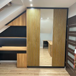 Cool-Ka MEBLE - Zabudowa pod skosem na poddaszu z biurkiem i szafą z lamelami oraz lustrem, widok na drugą część pomieszczenia.