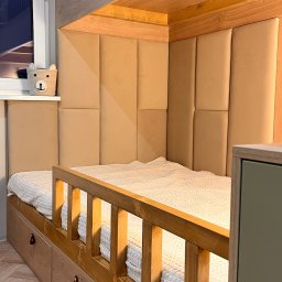 Cool-Ka MEBLE - Beżowe, tapicerowane panele ścienne tworzące zagłówek łóżka na antresoli z drewnianą barierką i szufladami, widok z dołu, obok okno z koszykiem na drobiazgi.