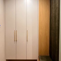 Cool-Ka MEBLE - Wnętrze przedpokoju z zabudową: gładkie, szare szafy z długimi, złotymi uchwytami, drewniany panel z pionowymi żłobieniami oraz tapicerowane siedzisko w odcieniach zieleni, całość na parkiecie...