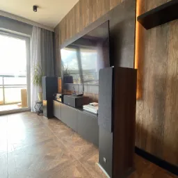 Nowoczesny salon z podświetlaną ścianą z imitacji drewna, szarą szafką RTV i systemem audio, widok na balkon przez przeszklone drzwi.