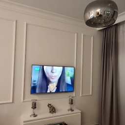 Dekorator-Gips Ewa Hładio - Elegancki salon z telewizorem na ścianie zdobionej dekoracyjnymi listwami, srebrna lampa wisząca, zasłony i firany w oknie.