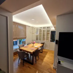 Jasne wnętrze jadalni z drewnianym stołem, szarymi krzesłami i oknem z roletą rzymską. Sufit podwieszany z oświetleniem LED tworzy nowoczesny klimat. Na stole wazon z różowymi kwiatami.