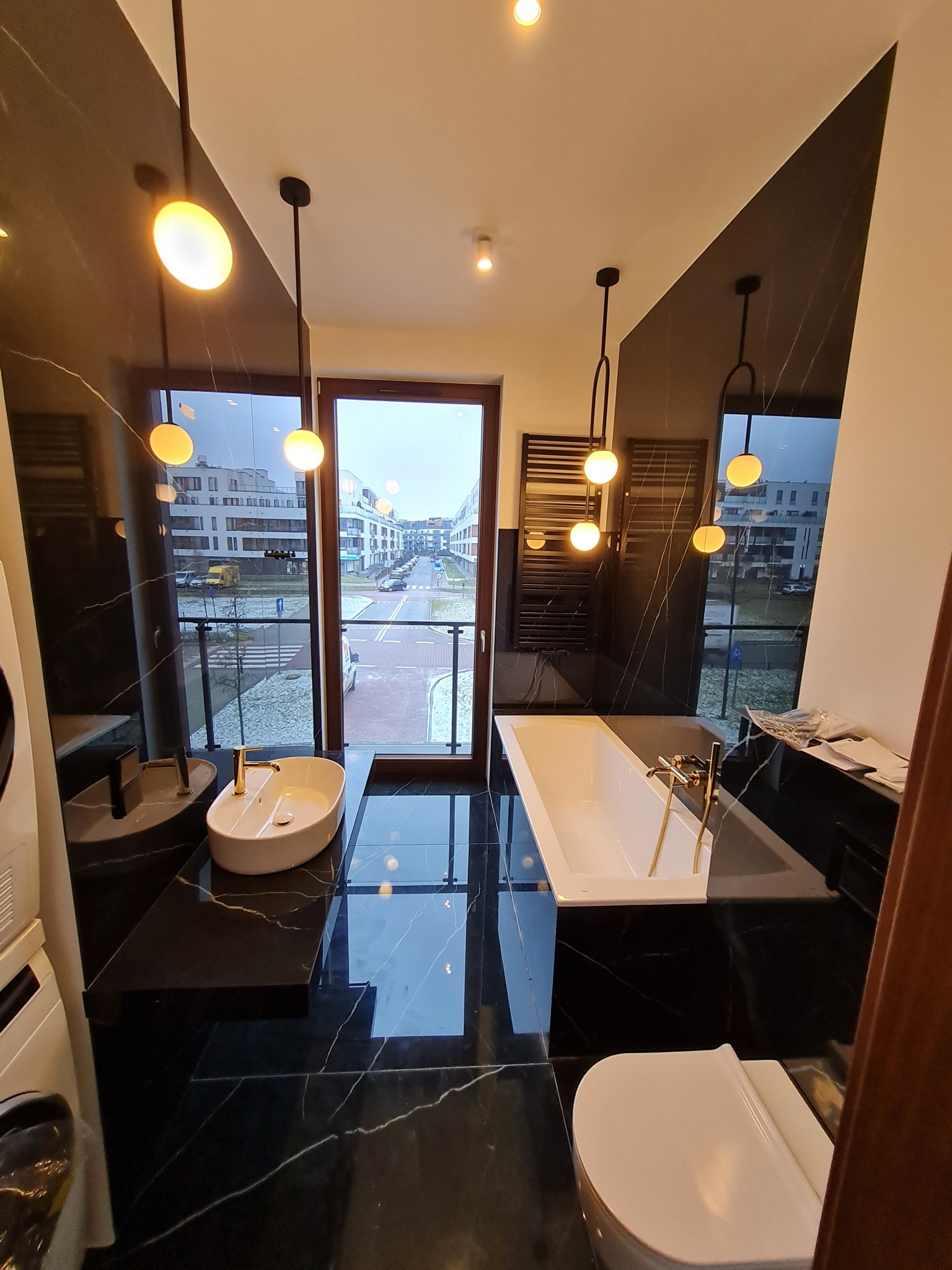 Nowoczesna łazienka z czarnymi marmurowymi płytkami, wiszącą umywalką i wanną, oświetlona kulistymi lampami, z widokiem na osiedle mieszkaniowe za oknem.