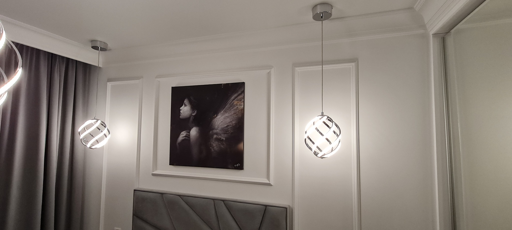 Eleganckie wnętrze sypialni z dwoma nowoczesnymi lampami wiszącymi o ażurowej konstrukcji, obrazem kobiety z anielskimi skrzydłami w ramie i szarym tapicerowanym zagłówkiem łóżka.
