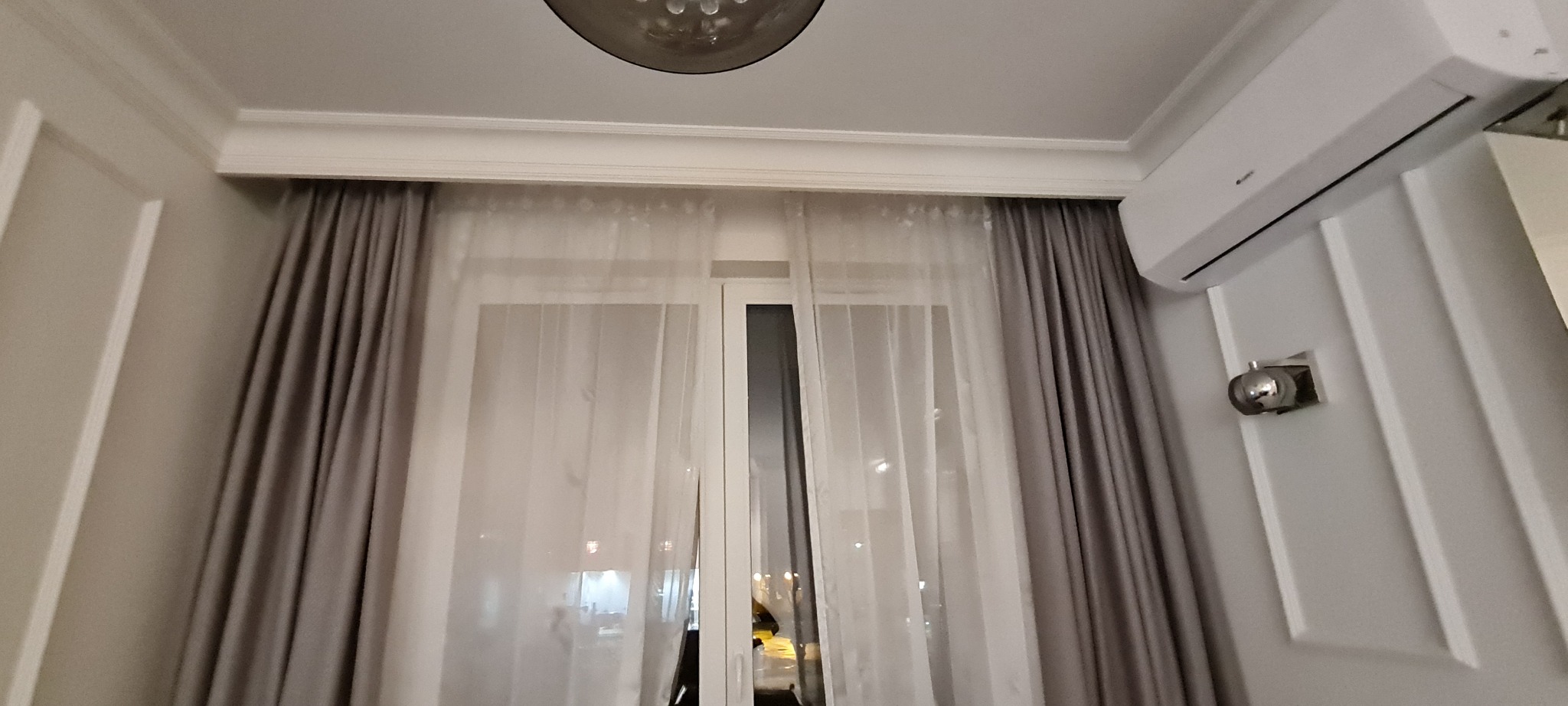 Wnętrze pokoju z oknem, zasłonami w odcieniach szarości i białymi firanami. Widoczny biały klimatyzator na ścianie z dekoracyjnymi listwami i okrągła lampa sufitowa.