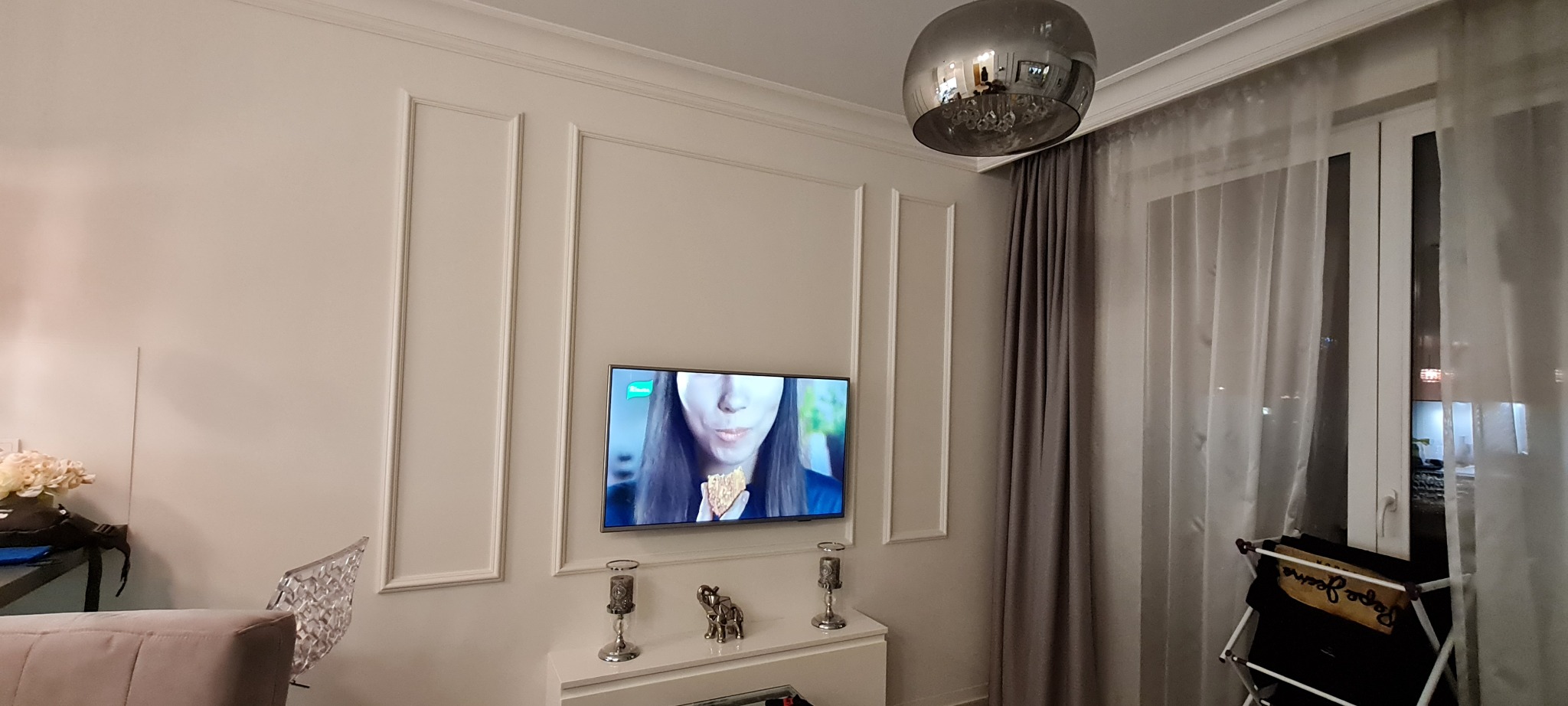 Elegancki salon z telewizorem na ścianie zdobionej dekoracyjnymi listwami, srebrna lampa wisząca, zasłony i firany w oknie.