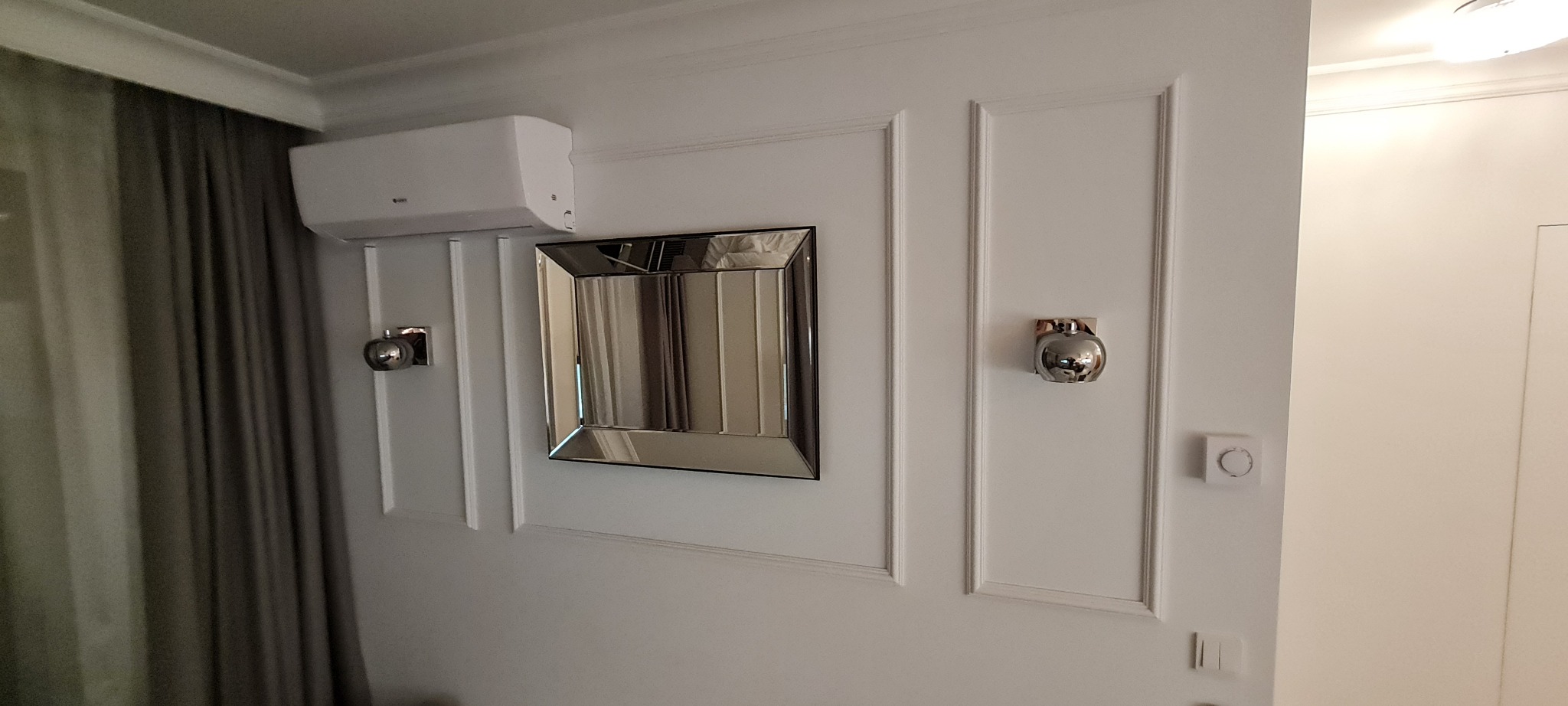 Biała ściana z dekoracyjnymi panelami, lustrem w srebrnej ramie, klimatyzatorem i dwoma chromowanymi lampami w kształcie jabłek.