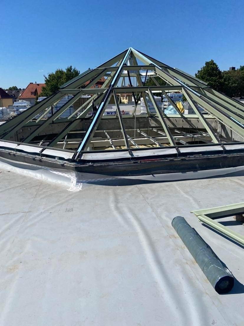 Montaż szkieletu piramidalnego świetlika dachowego na płaskim dachu pokrytym membraną, widoczne elementy konstrukcyjne i rolka materiału izolacyjnego.
