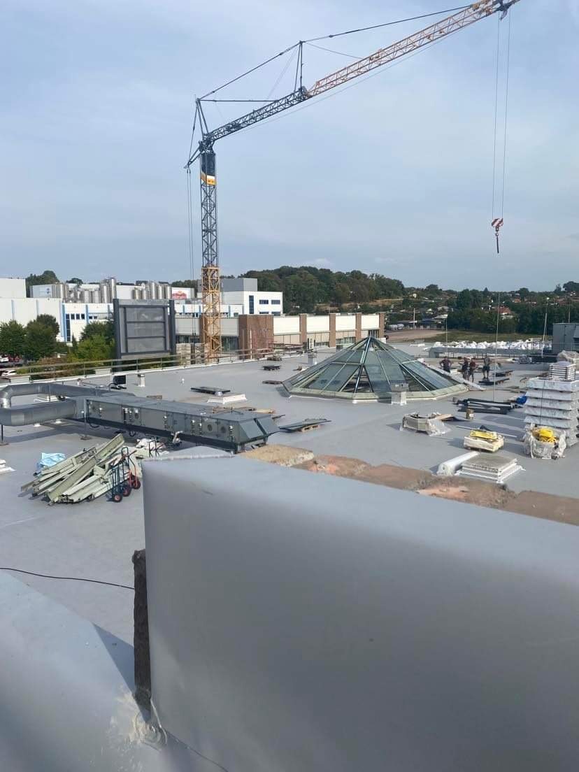 Widok z góry na rozległy płaski dach budynku przemysłowego z przeszkloną piramidą, z widocznym dźwigiem budowlanym w tle i elementami wentylacji na dachu.